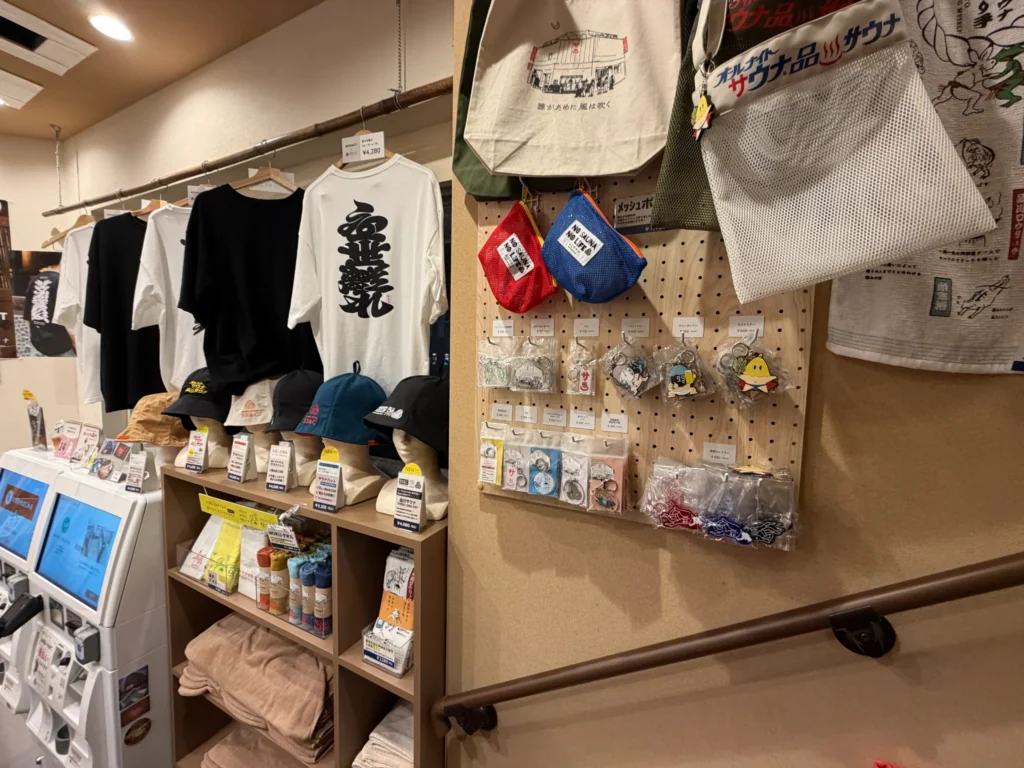 品川サウナのオリジナルグッズ販売コーナー。壁面にTシャツやトートバッグが掛けられ、棚にはサウナハットやキャップ、ステッカーなどの小物が並んでいる。有孔ボードにはキーホルダーやアクセサリーが陳列されている