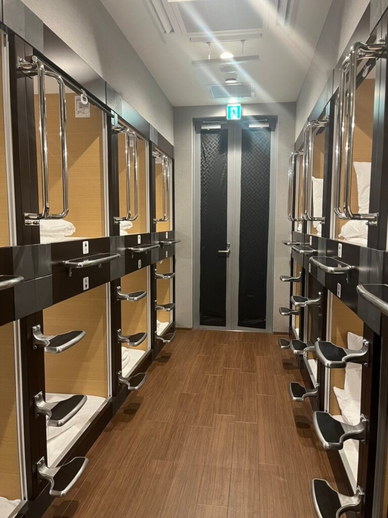 品川サウナのカプセルホテルフロア通路。左右に2段構成のカプセルユニットが並び、各カプセルには白いタオルが用意されている。木目調の床とクロームメッキの金属フレームで清潔感のある空間