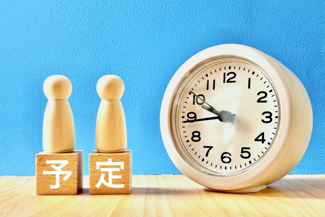 アナログ目覚まし時計と「予定」と書かれた木製ブロック(水色背景のイメージ写真)