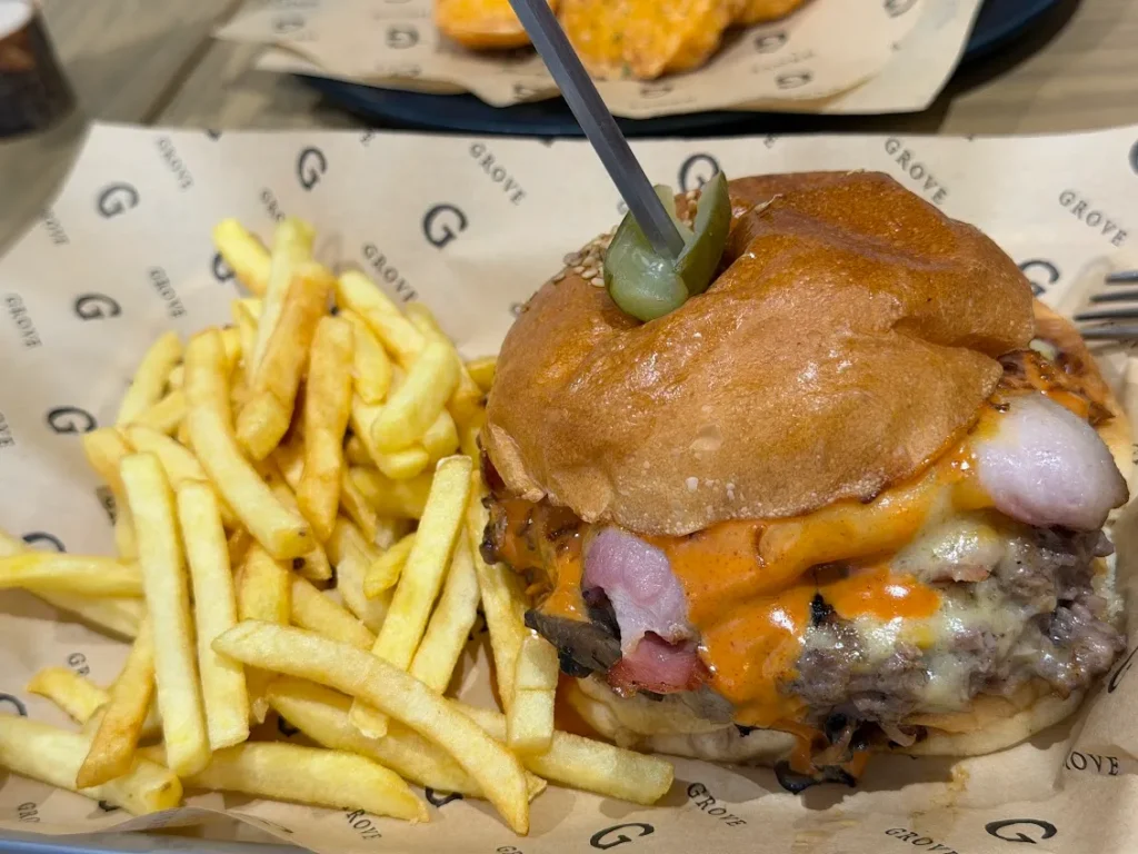 GROVEのロゴが並ぶ包み紙の上に置かれたハンバーガー。つやのあるバンズに、とろけたチーズと肉厚のパティ、ピックで刺したオリーブがのり、横にはたっぷりのフライドポテトが添えられている。