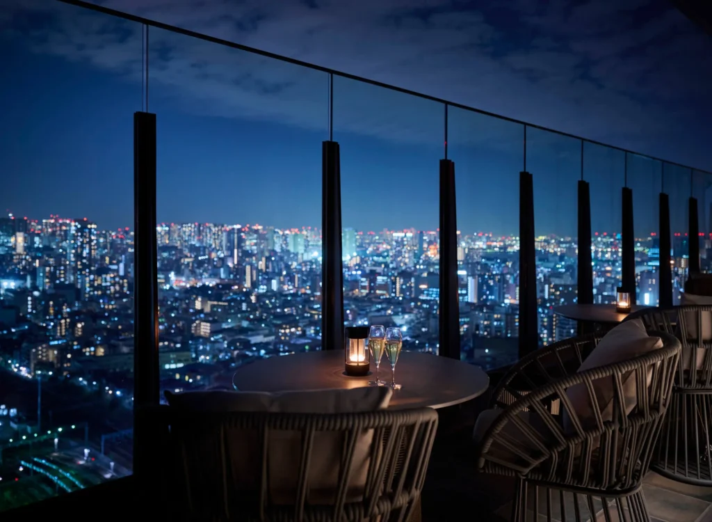 ルーフトップバーのテラス席から見た東京の夜景。ガラスの手すり越しに無数の街明かりが地平線まで広がり、手前の丸テーブルにはランタン型のキャンドルとグラス、籐製のアームチェアが並ぶ。空は群青色の夕暮れ。