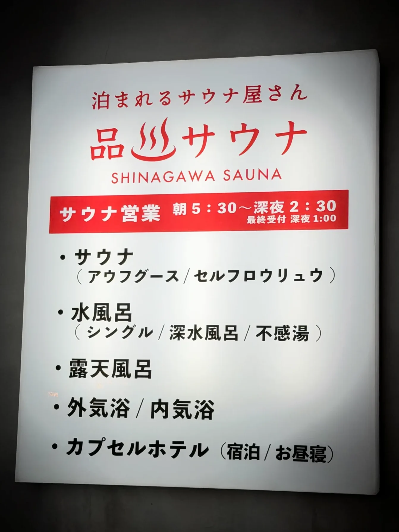 品川サウナの外看板。「泊まれるサウナ屋さん 品川サウナ」のロゴと営業時間（朝5:30〜深夜2:30）が記載されており、サウナ（アウフグース／セルフロウリュウ）、水風呂（シングル／深水風呂／不感湯）、露天風呂、外気浴／内気浴、カプセルホテル（宿泊／お昼寝）の施設一覧が掲示されている