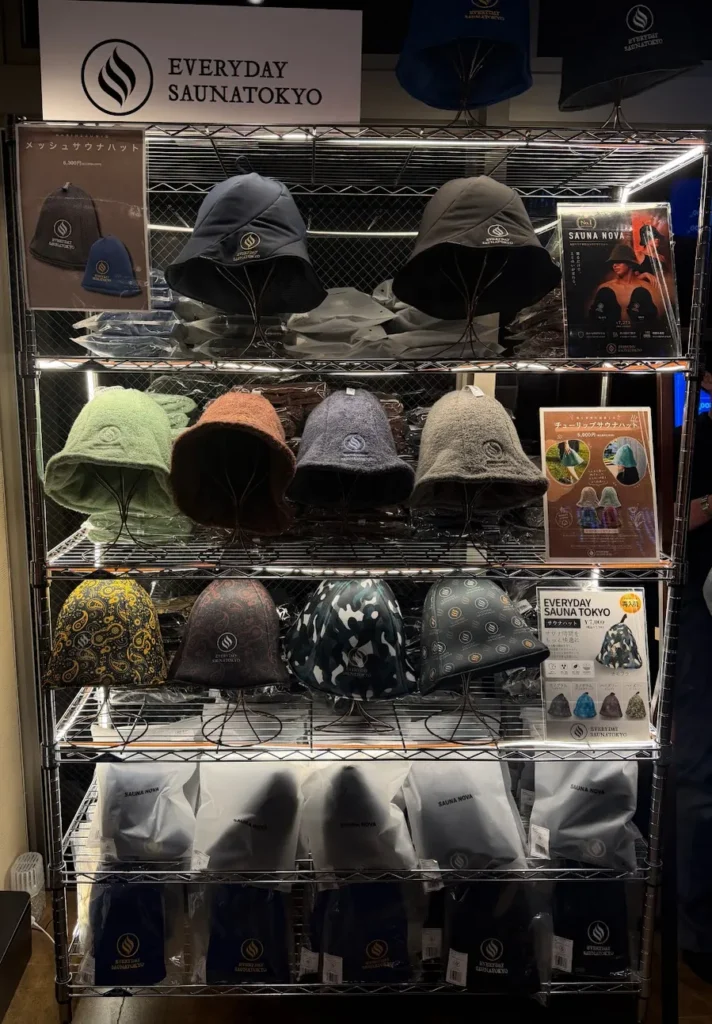 オリジナルグッズのサウナハット陳列棚。金属ラックに各色のサウナハットが並ぶ（青・黒のキャップ、グレー・緑・ピンクのサウナハット、花柄・迷彩柄のもの）。EVERYDAY SAUNATOKYOのロゴ看板と商品ポップ