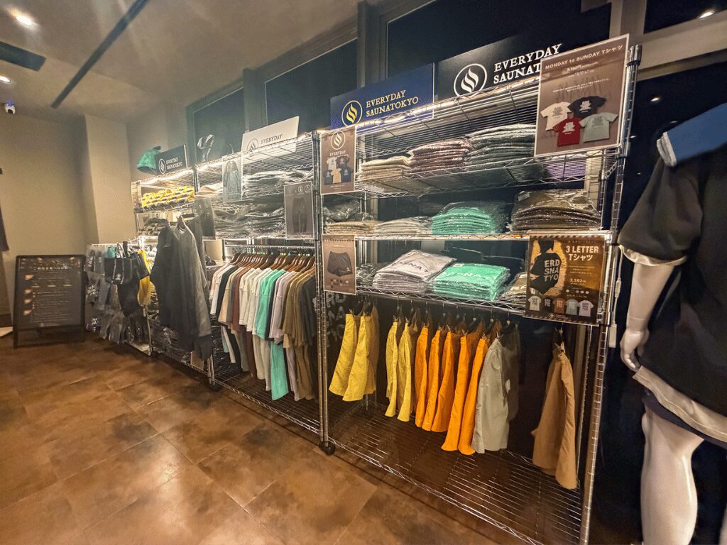 オリジナルグッズ販売コーナー。金属製ラックにTシャツやパーカーがハンガーで掛けられている（黄色、グレー、白など複数色）。上段にたたまれた衣類やパッケージ商品。EVERYDAY SAUNATOKYOのロゴ看板。木目調の床