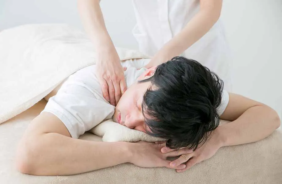 白い施術室でうつ伏せに横たわる男性の背中を、白い制服を着た施術者がマッサージしている。ベージュのタオルが敷かれたベッドの上で、明るく清潔感のある空間