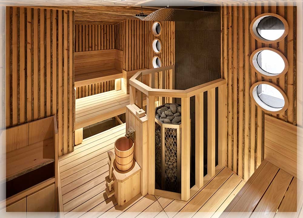 ケロサウナ内部。縦板張りの木壁に囲まれたコンパクトな空間。中央に円柱型のサウナストーブ（サウナストーンを積載）が木製柵で囲まれ、横に木製バケツが置かれている。右壁に3つの丸窓、左奥に2段ベンチとLED照明が見える
