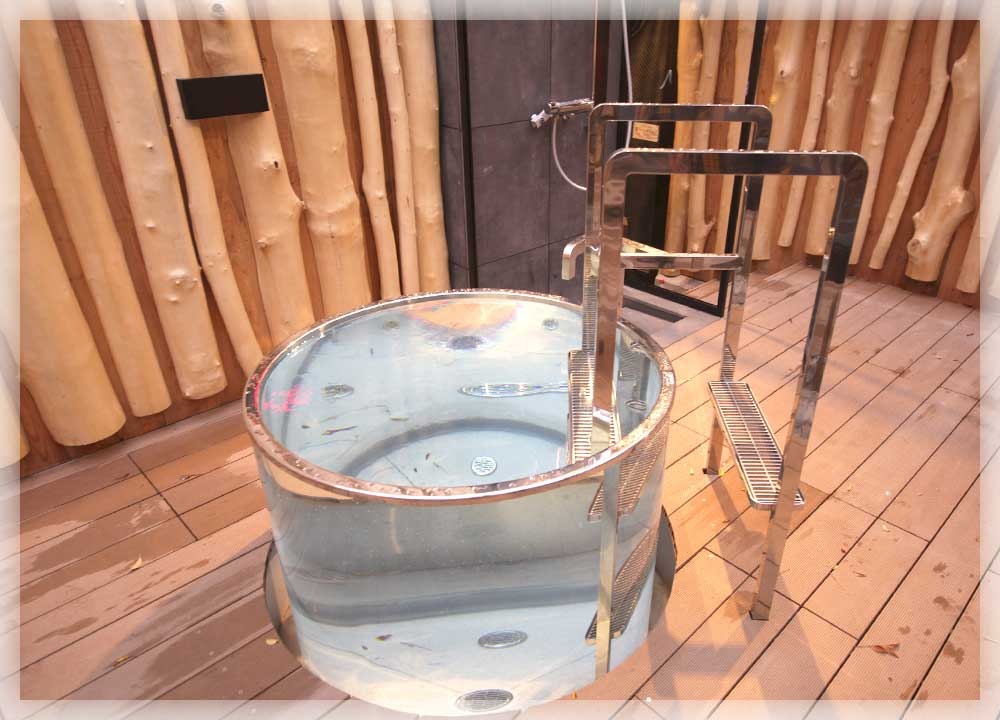 屋外ウッドデッキの上に置かれた一人用の円形金属浴槽（ガルバナイズドスチール製）に水が張られている。横にステンレスのステップ梯子。背景に丸太を縦に並べた木製フェンスが立つ