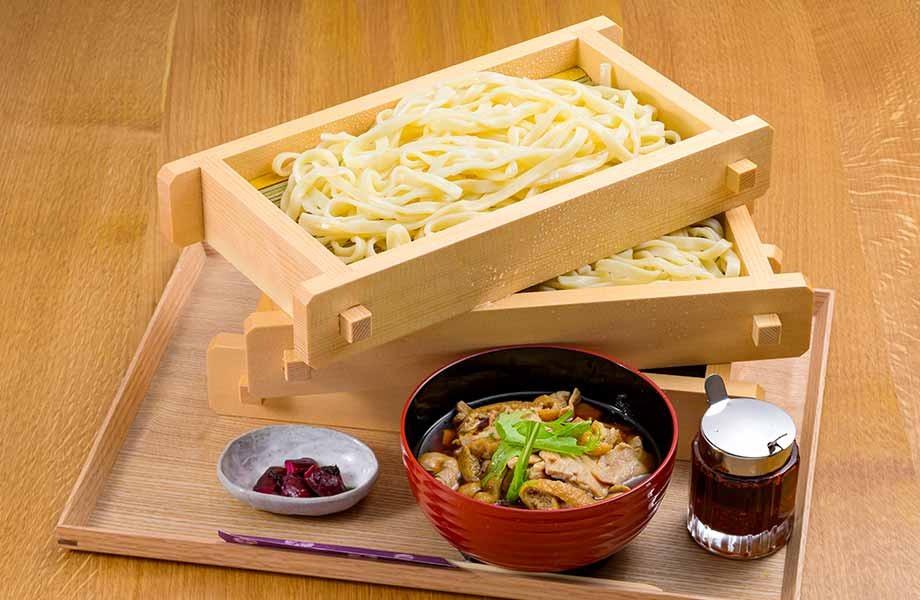 木製トレイに盛り付けられた肉汁うどんセット。木箱に入ったうどん麺、赤い漆器の椀に肉と野菜の付けつゆ、小皿の梅干し、グラスのつゆが並ぶ。箸が添えられている