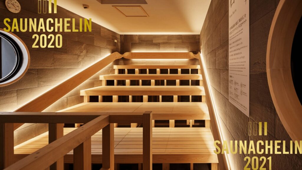 SAUNACHELIN 2020第2位・2021第3位の金色ロゴが表示された、かるまる池袋の岩サウナ内部。多段式の木製ベンチにLED間接照明が灯り、左壁に丸窓、右壁に石材パネルと案内板が見える
