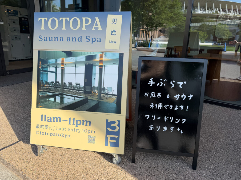 施設入口に置かれた案内サインボード。「TOTOPA Sauna and Spa 男性 Men」「11am–11pm 最終受付10pm」「3F」と書かれ、隣の黒板に『手ぶらでお風呂&サウナ利用できます!フリードリンクあります』と手書きされている