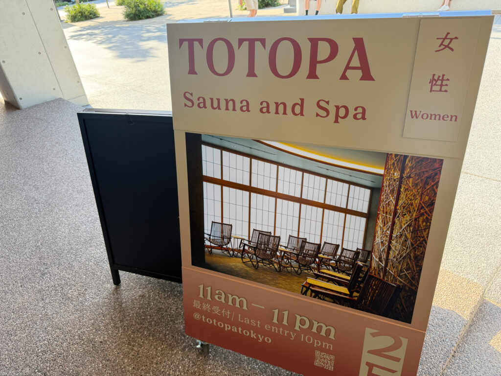施設入口に置かれた案内サインボード。「TOTOPA Sauna and Spa 女性 Women」「11am–11pm」「2F」と書かれ、ラウンジの写真が添えられている