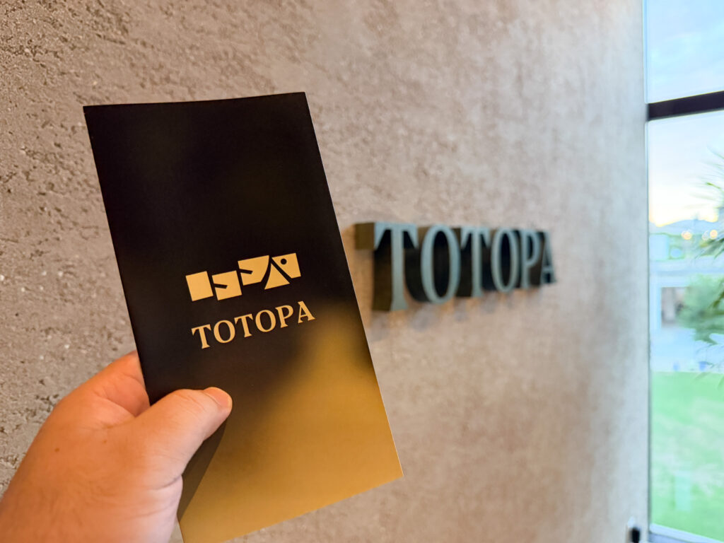 手に持った黒い「TOTOPA」のガイド冊子と、その奥の壁に取り付けられたメタリックな「TOTOPA」のロゴサイン