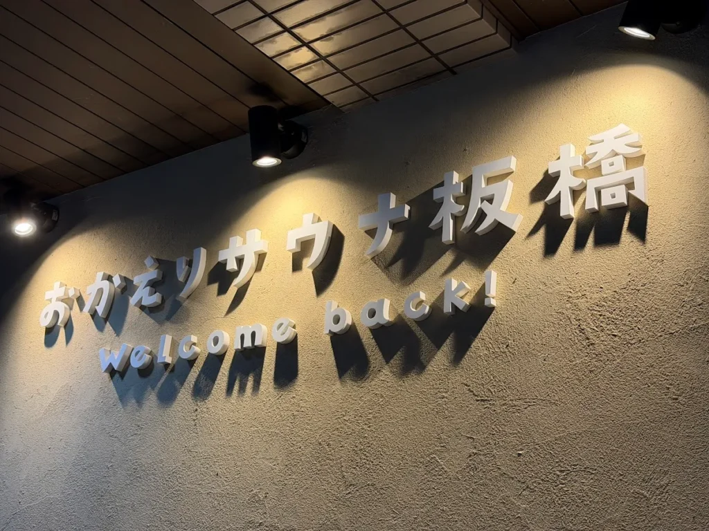 おかえりサウナ板橋の館内。暗い壁面に白い筆文字で「おかえりサウナ板橋」のロゴサインが掲げられ、その下に小さくwelcome back!と英字表記がある