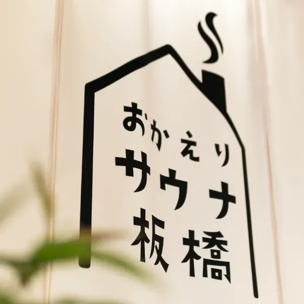 おかえりサウナ板橋のロゴ看板。家の形のシルエットの中に「おかえりサウナ板橋」と筆文字で書かれた暖簾と、手前にのぞく観葉植物の緑